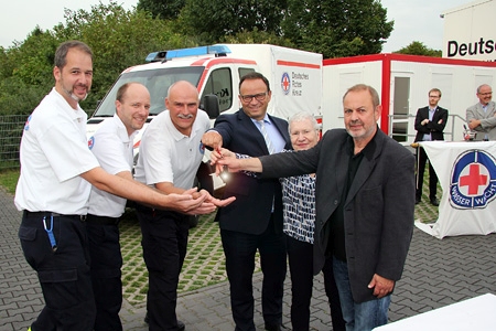 Jan Höpfner, Christian Vorstius und Klaus Püttmann von der Wasserwacht nehmen den Schlüssel des neuen Gerätewagens von Sparkassenvorstand Frank-Rainer Laake, Gisela Bangen, Vorsitzende des DRK-Kreisverbandes Niederrhein, und Ulrich Rassier, Koordinator Gefahrenabwehr für den Kreis Wesel, entgegen. Im Hintergrund beobachten Pfarrer Udo Otten und Pastoralreferent Werner Koschinski die Schlüsselübergabe für das zuvor von ihnen gesegnete Fahrzeug.