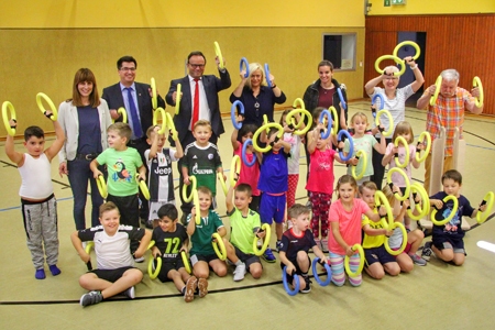 Die Kinder der Klasse 1 b schwingen ihre neuen Smovey-Ringe, die Turnhalle der Städtischen Gemeinschaftsgrundschule brummt. Auch die stellvertretende Schulleiterin Petra Engeln (v.l.n.r.), Bürgermeister Frank Tatzel, Sparkassenvorstand Frank-Rainer Laake, Helga Karl vom städtischen Fachbereich Schule und Sport, Studienpraktikantin Charlyn Meyer sowie die Bewegungsaktivisten Marianne und Detlev Friedriszik brummen schwungvoll mit.