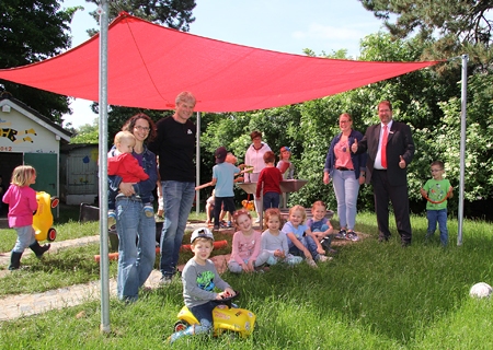 Barbara Kellings mit Sohn Jakob und Jörg Rösken vom Förderverein (links), Christel Spitz-Güdden von der Verbundleitung der Kirchengemeinde (hinten an der Wasserpumpe), Kita-Leiterin Elisa Hemmers-van Husen und Volker Oppers von der Sparkasse am Niederrhein (rechts) freuen sich über das neue Sonnensegel, das die Kinder der Kita St. Michael beim Spielen schützt.