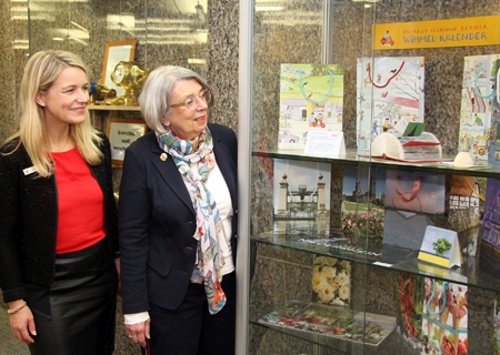 Geschäftsstellenleiterin Katrin Steffans und Hobbykünstlerin Monika Paaßen (r.) stehen an der Vitrine mit den aus Kunstkalender-Blättern gefalteten Tragetaschen.