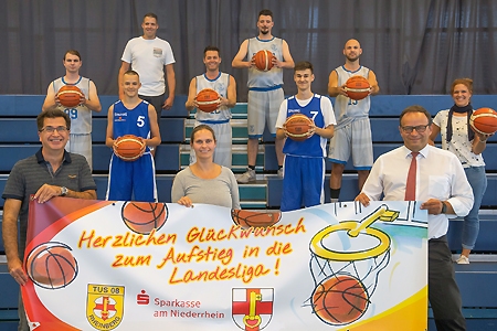 Bürgermeister Frank Tatzel, Stefanie Kaleita (stellv. Leiterin des Fachbereichs Schule-Sport-Kultur) 	und Sparkassendirektor Frank-Rainer Laake präsentieren das Aufstiegsbanner. Dahinter steht die erfolgreiche Herrenmannschaft des TuS 08 mit Trainerin Anika Illbruck-Schuurmann (r.). Die Sparkasse am Niederrhein spendete 1.500 Euro für die Aus- und Fortbildung der Übungsleiter in der Basketball-Abteilung.